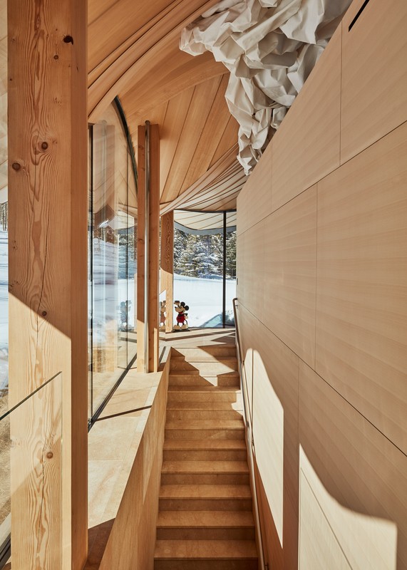 Interior view of a wooden staircase in a modern house with sunlight filtering through a window overlooking a snow-covered landscape.