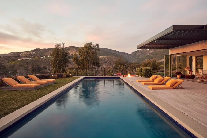 An outdoor swimming pool is framed by lounge chairs on the grass and wood, with a mountain range visible at dusk.