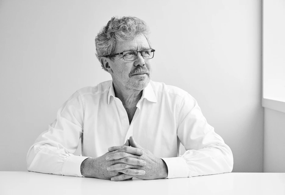 Black and white portrait of a mature adult male with glasses, white shirt, seated at a table against a neutral background in a bright indoor space.