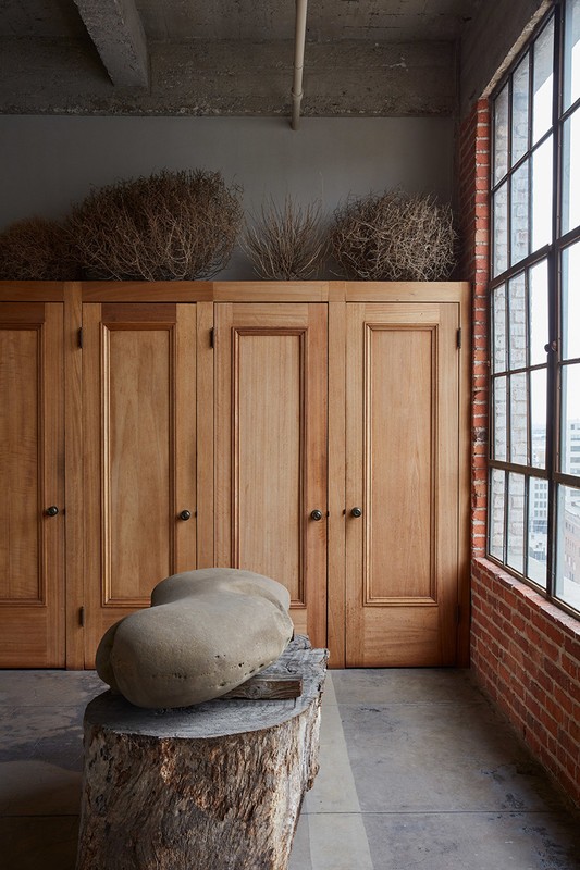 Interior shot of sculpture on a wooden stump in front of wooden cabinets, topped with tumbleweeds in a room with a brick wall.