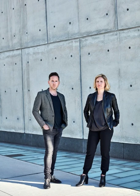 Full shot of a confident adult man and woman standing side-by-side in an urban setting, wearing blazers and dark clothing against a concrete wall.