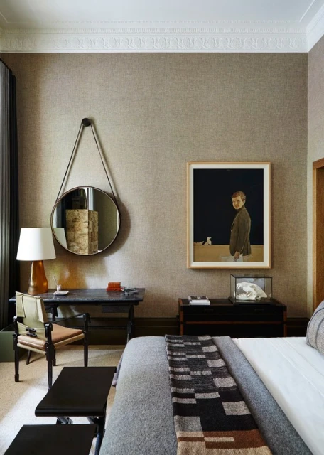 Bedroom interior with a bed, desk, artwork, round mirror, and neutral wallpaper, creating a modern and stylish living space.