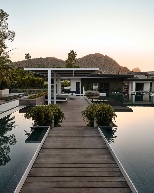 Exterior view of modern home with infinity pool, patio cover, landscaping, palm trees, and mountain in background during sunset.