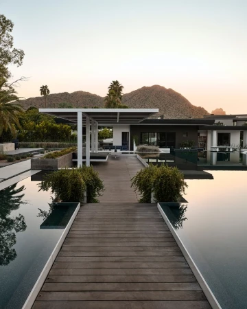 Exterior view of modern home with infinity pool, patio cover, landscaping, palm trees, and mountain in background during sunset.