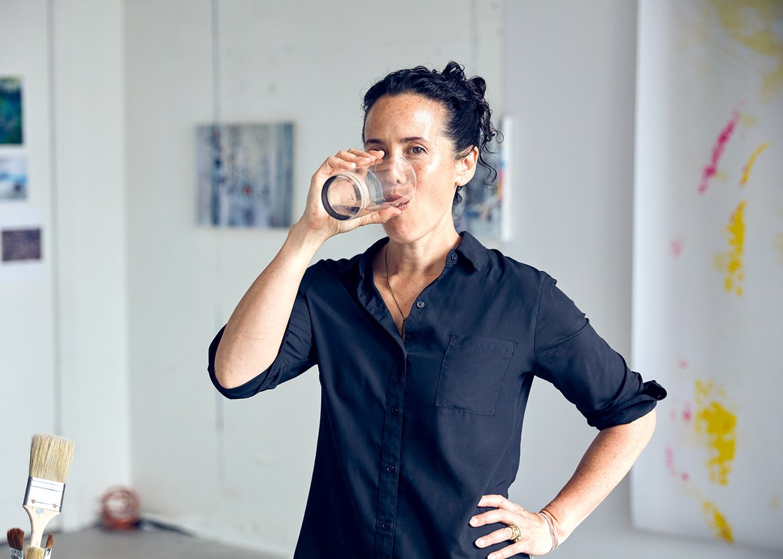 Adult woman drinking water from clear glass, standing in bright art studio, wearing a dark button-up shirt with art and paintings visible.
