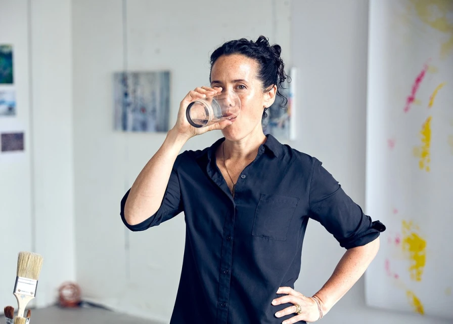 Adult woman drinking water from clear glass, standing in bright art studio, wearing a dark button-up shirt with art and paintings visible.