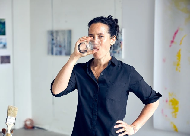 Adult woman drinking water from clear glass, standing in bright art studio, wearing a dark button-up shirt with art and paintings visible.