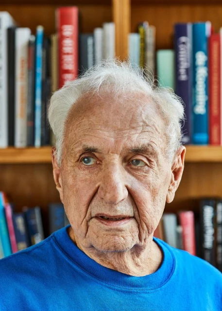 Close-up portrait of an elderly adult male with white hair wearing a blue shirt, set against a background of a wooden bookshelf filled with books.