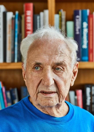 Close-up portrait of an elderly adult male with white hair wearing a blue shirt, set against a background of a wooden bookshelf filled with books.