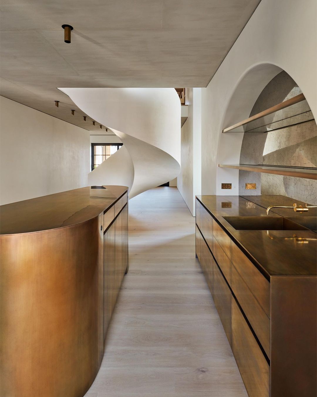 Interior design featuring a spiral staircase and kitchen area with light wood floors, brass-colored cabinetry, and white walls, creating a modern aesthetic.