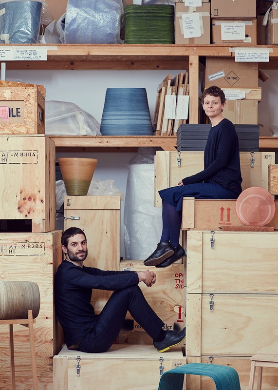 A man and a woman sit surrounded by wooden crates and shelves filled with pottery, vases, and boxes inside an industrial space.