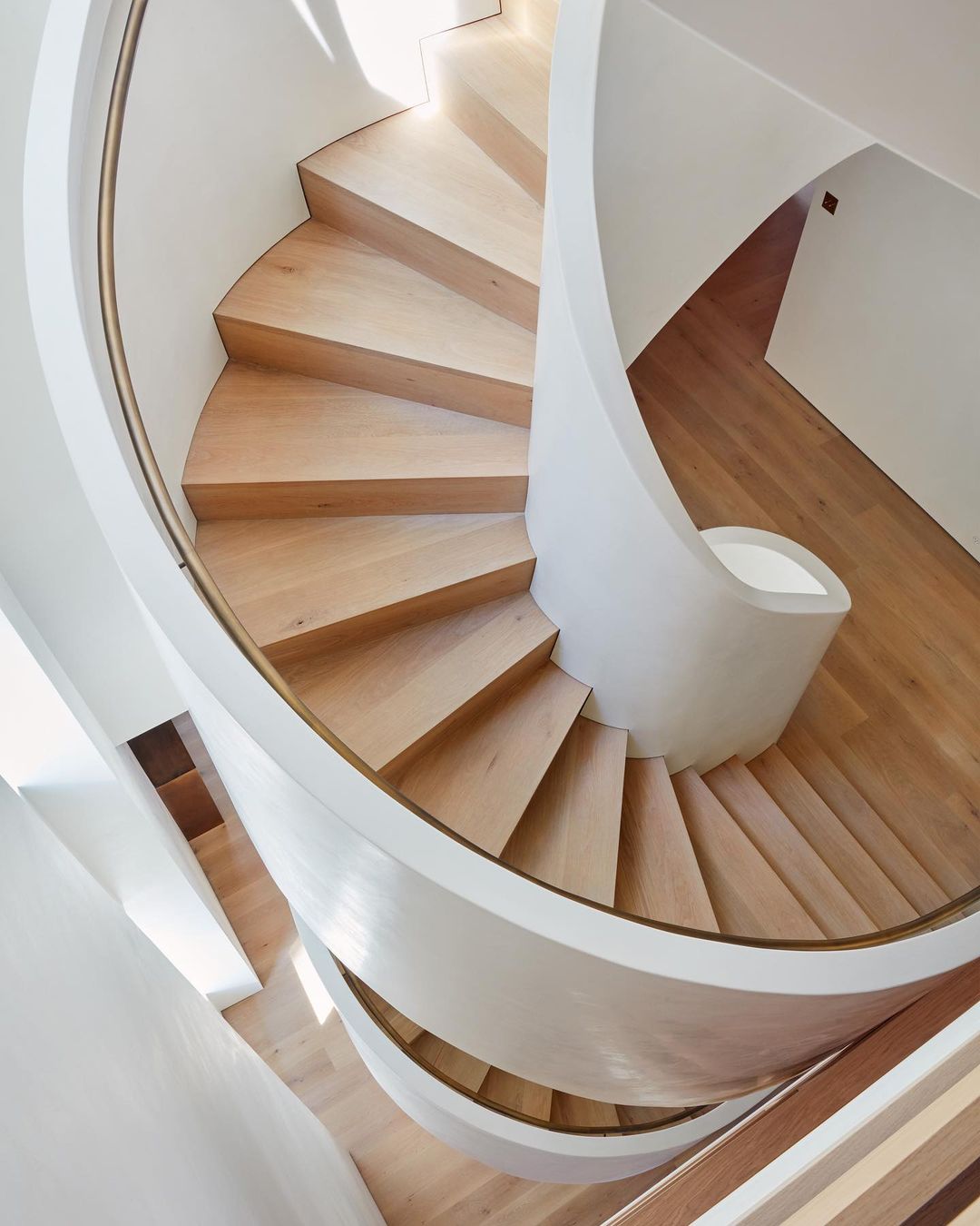 An upward view of a modern spiral staircase in an indoor architectural space, featuring wooden steps and white curved walls, bathed in natural light.