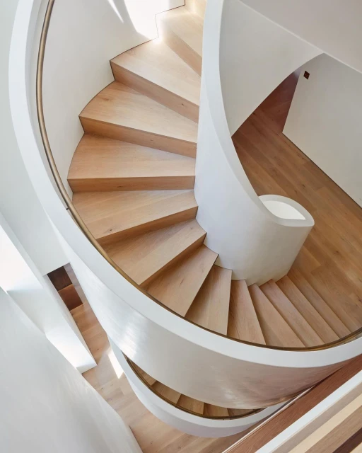 An upward view of a modern spiral staircase in an indoor architectural space, featuring wooden steps and white curved walls, bathed in natural light.