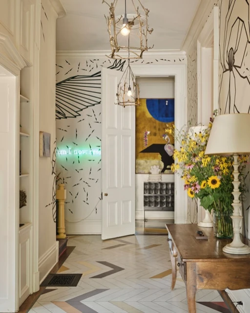 Interior view of a hallway featuring a herringbone tile floor, table with flowers, unique light fixtures, and artistic wallpaper.