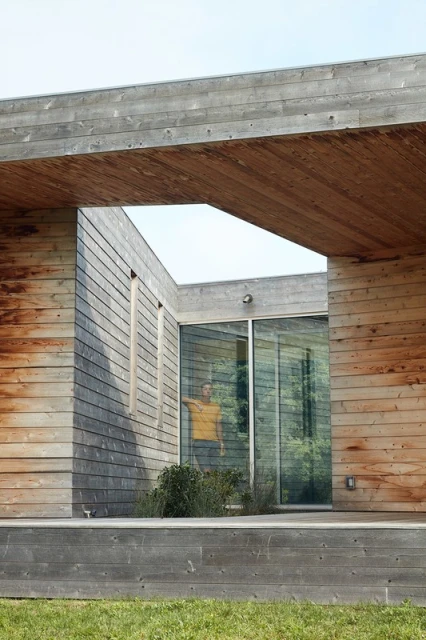 Modern architecture featuring a wood-clad building with a person visible through the glass, surrounded by greenery and a serene atmosphere.