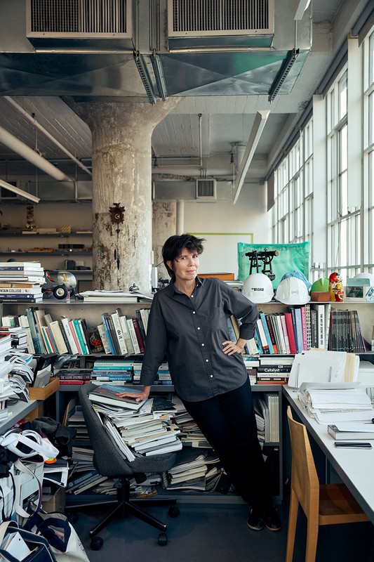 Adult female poses in a cluttered office with bookshelves, papers, and hard hats near a large window with natural lighting.