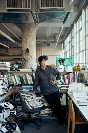 Adult female poses in a cluttered office with bookshelves, papers, and hard hats near a large window with natural lighting.
