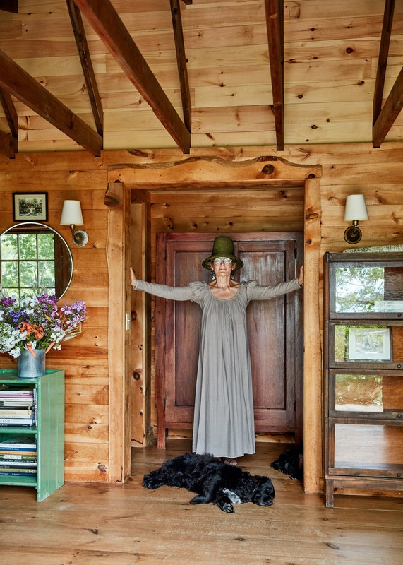 Woman in a long dress and hat stands in a doorway of a wood cabin with two black dogs laying on the wood floor.