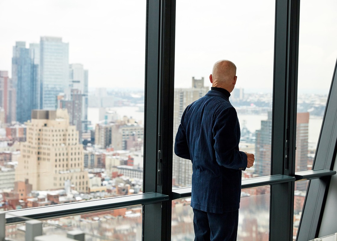 Back view of a bald adult man in a navy jacket looking out a large window at a cityscape with skyscrapers and gray skies on an overcast day.