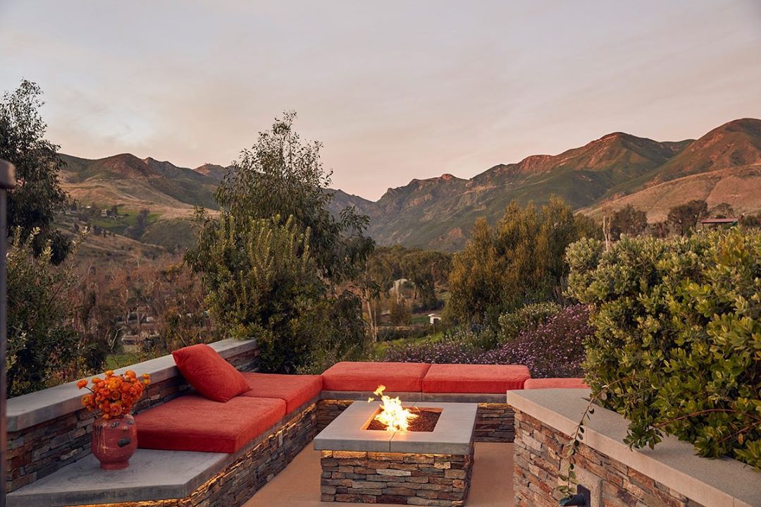 Outdoor patio area with orange cushions, stone fire pit with flames, potted flowers, and a view of grassy mountains in the background.