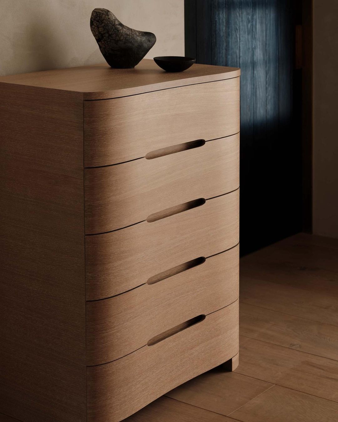 Interior design featuring a light wood dresser with drawers, topped with a stone sculpture and small bowl, in a dimly lit room.