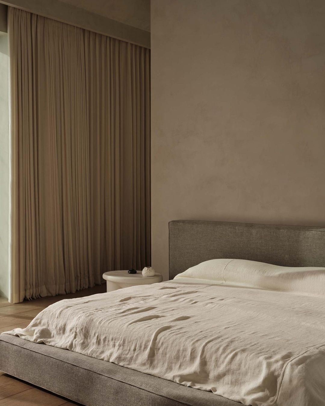 Bedroom interior featuring a bed with neutral bedding, a headboard, and a bedside table against a beige wall and draped curtains.