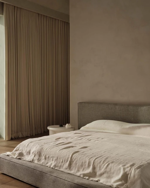 Bedroom interior featuring a bed with neutral bedding, a headboard, and a bedside table against a beige wall and draped curtains.