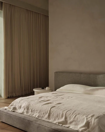Bedroom interior featuring a bed with neutral bedding, a headboard, and a bedside table against a beige wall and draped curtains.