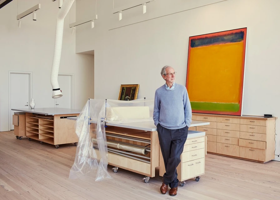 An elderly man stands leaning against a storage unit in a light-filled room, with abstract art and tables visible, creating a serene and artistic scene.