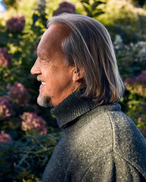 Profile portrait of mature adult man with gray hair and beard in a gray turtleneck sweater, outdoors in a natural setting with blooming bushes.