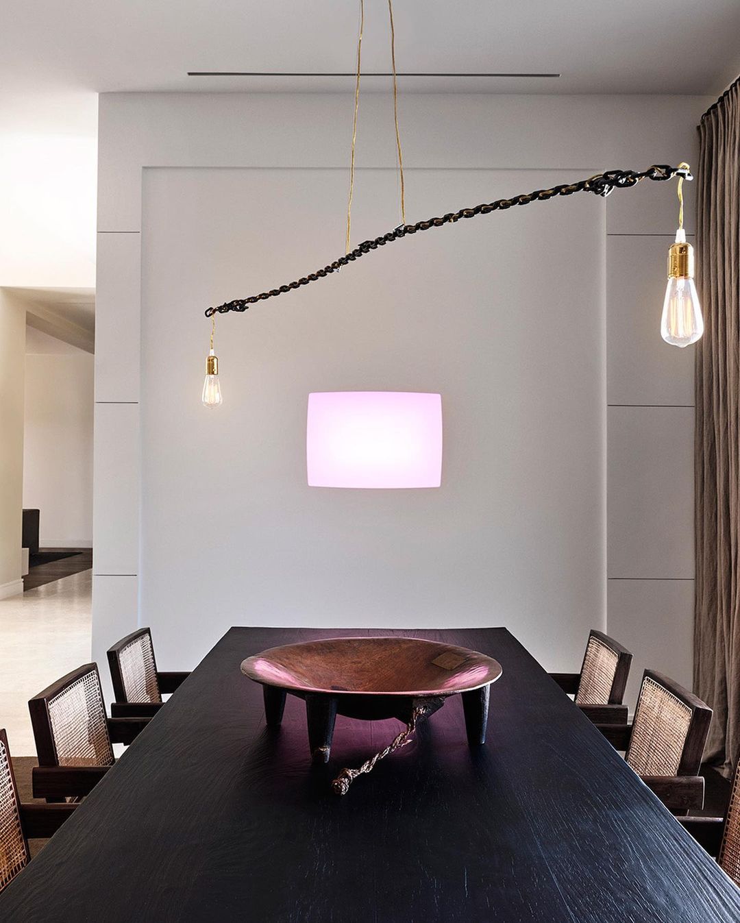 Dining room interior featuring a black wooden table, straw-backed chairs, and a unique chain-linked light fixture with Edison bulbs.
