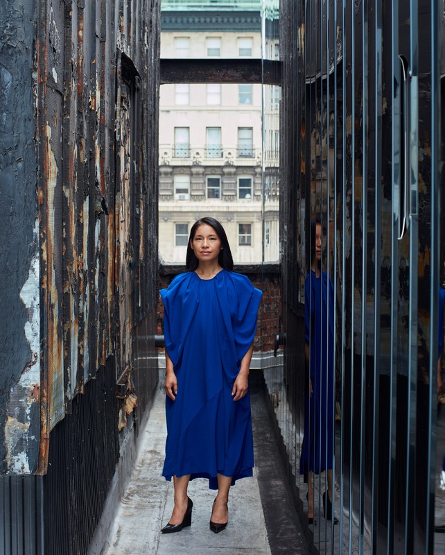 A woman in a blue dress stands in an urban alleyway framed by weathered walls. A reflection in the background mirrors her image.
