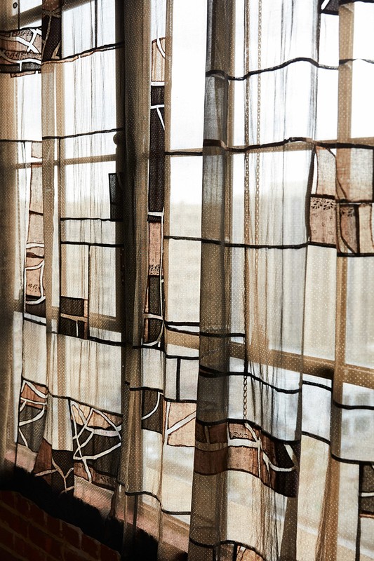 Sheer curtains with a geometric design hang in front of a window. The colors are neutral tans, whites, and browns, and black in an indoor setting.