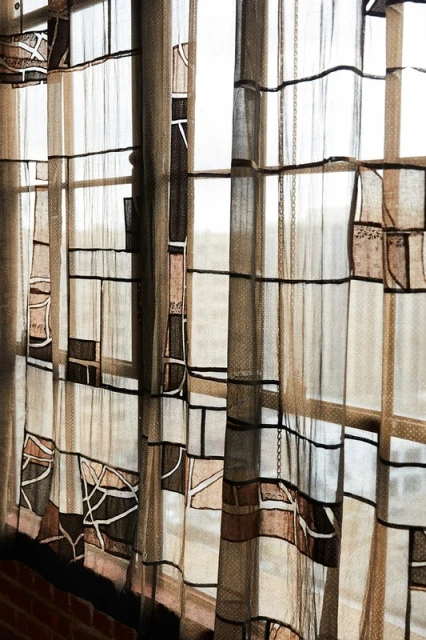 Sheer curtains with a geometric design hang in front of a window. The colors are neutral tans, whites, and browns, and black in an indoor setting.