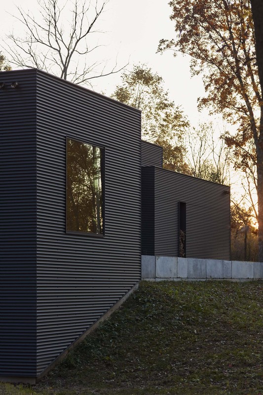 Exterior of a modern black corrugated metal building with rectangular windows, set in a grassy landscape with trees in the background.