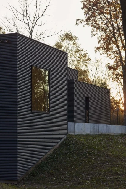 Exterior of a modern black corrugated metal building with rectangular windows, set in a grassy landscape with trees in the background.