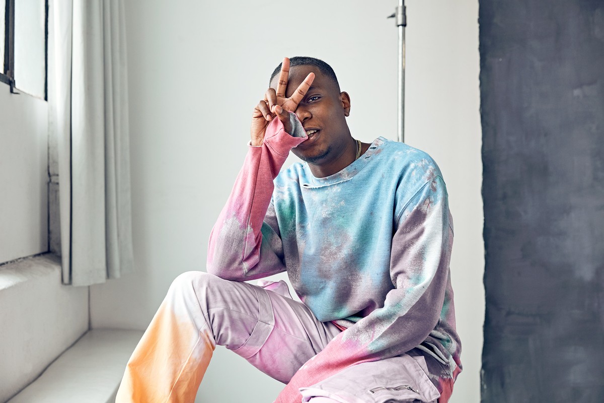 Man in tie-dye top gives peace sign while seated in a photo studio with white walls and a gray accent panel, bathed in natural light.