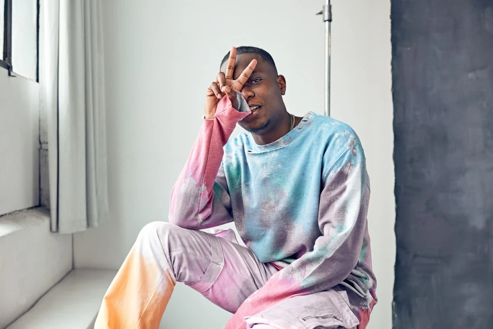 Man in tie-dye top gives peace sign while seated in a photo studio with white walls and a gray accent panel, bathed in natural light.