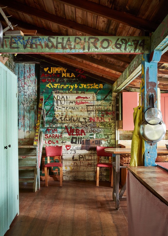 View of rustic interior with painted wood walls and ceiling, red chairs, round table, and hanging pots and pans in a rural setting.