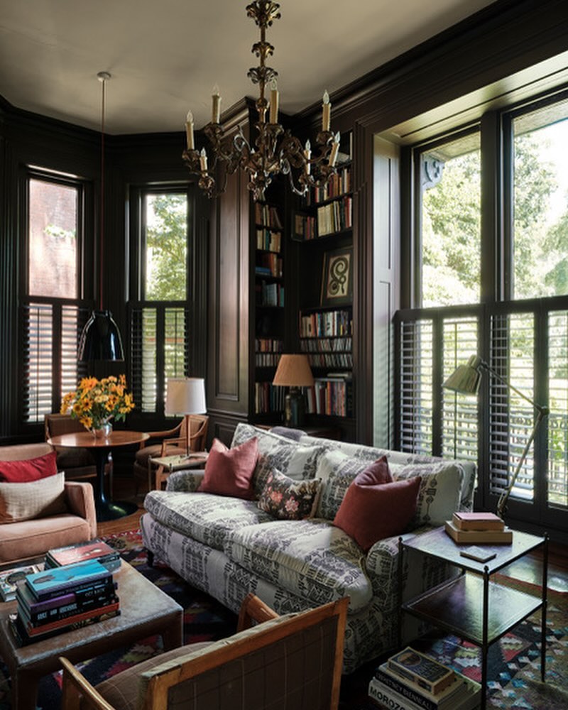 Cozy living room interior with patterned sofa, bookshelves, chandelier, and natural light streaming through window with shutters.