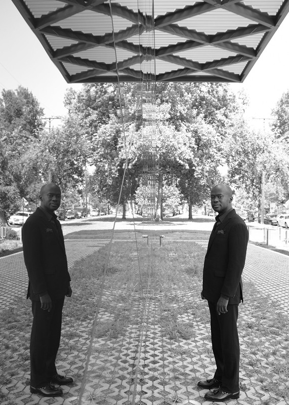 Black and white portrait of two adult males in suits standing outdoors with symmetrical background of trees and architecture.