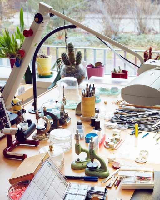 Watchmaker's workbench with tools, watch parts, and equipment under a lamp, near a window, in a brightly lit indoor environment.