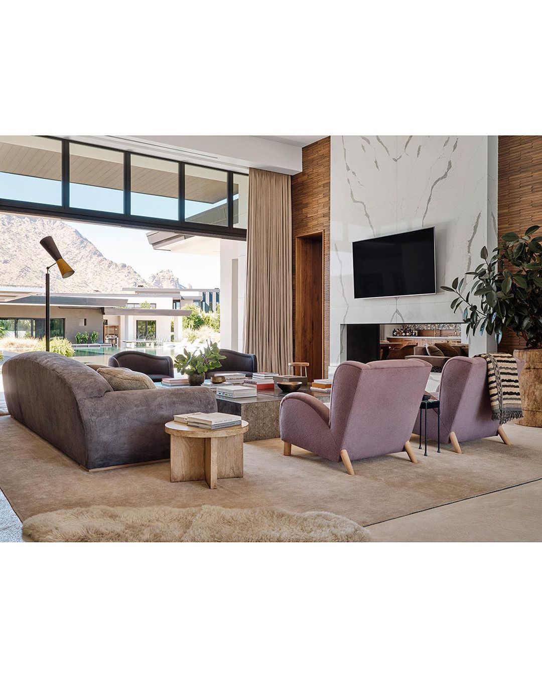 Living room interior showcasing a gray sofa, two pink armchairs, and a marble fireplace with a mounted TV in a modern home setting.