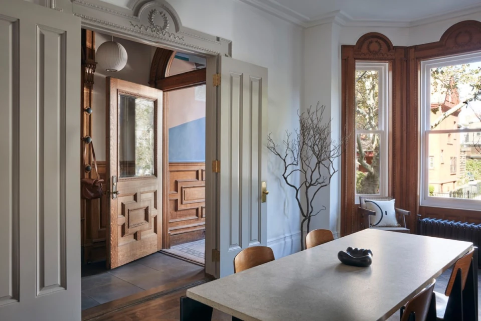 Interior view of a dining room with a large concrete table, wood chairs, a faux tree, and open doors leading to a hallway and entryway.