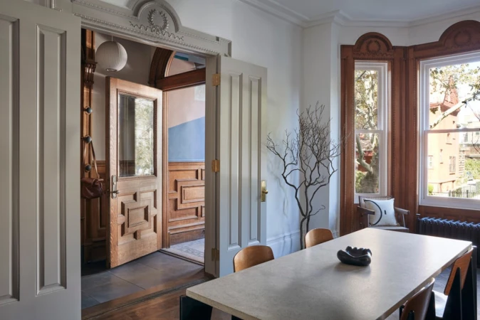 Interior view of a dining room with a large concrete table, wood chairs, a faux tree, and open doors leading to a hallway and entryway.