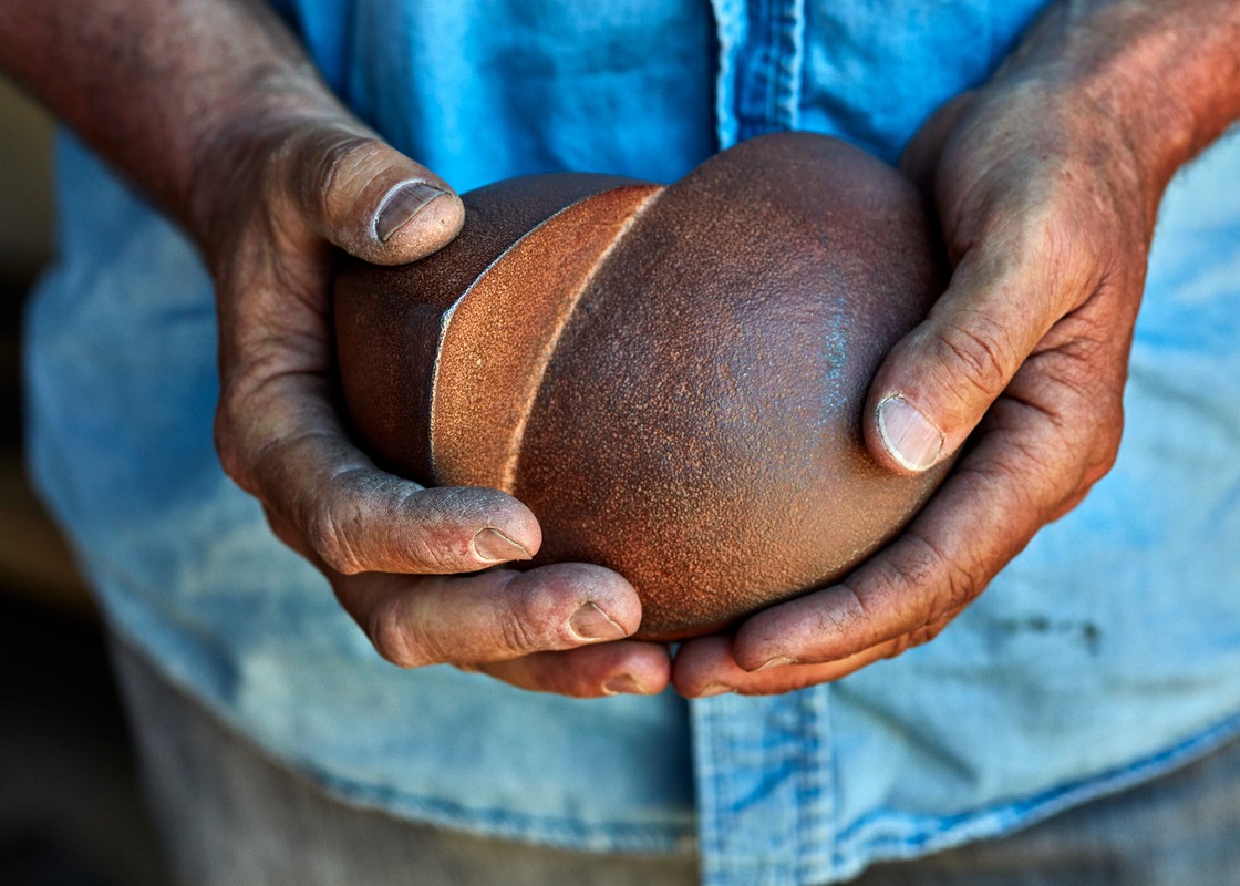 Hands holding a crafted brown sphere with a lighter-colored indentation. The person wears a light blue shirt.