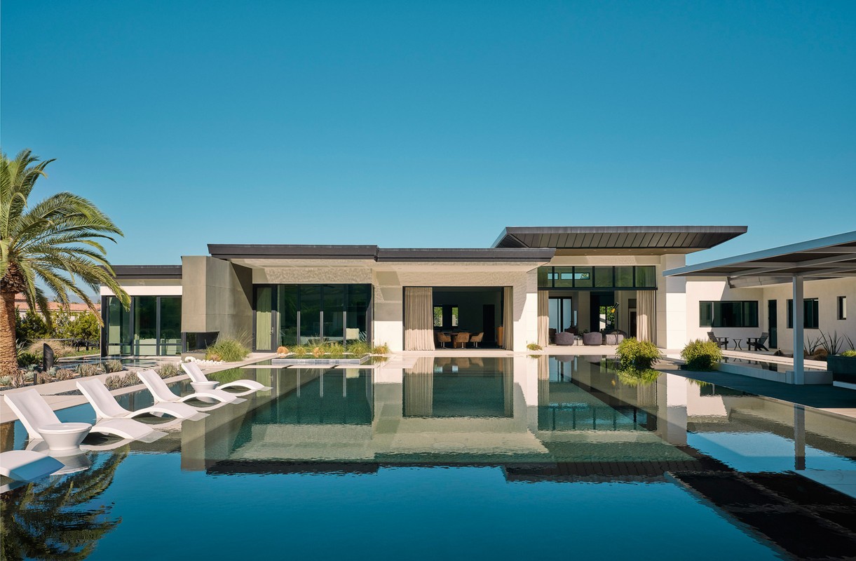 Exterior view of a modern house with a dark swimming pool, white lounge chairs, and a palm tree against a clear blue sky.