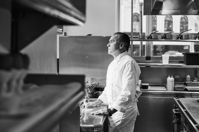 Black and white shot of a chef in a white coat standing in a professional kitchen with stainless steel appliances.