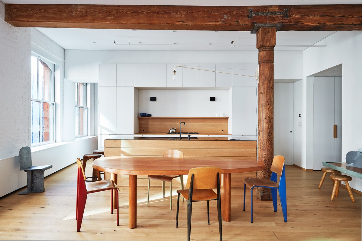 Bright, modern interior with an oval dining table surrounded by colorful chairs, set against a backdrop of white cabinets and rustic wooden beams.