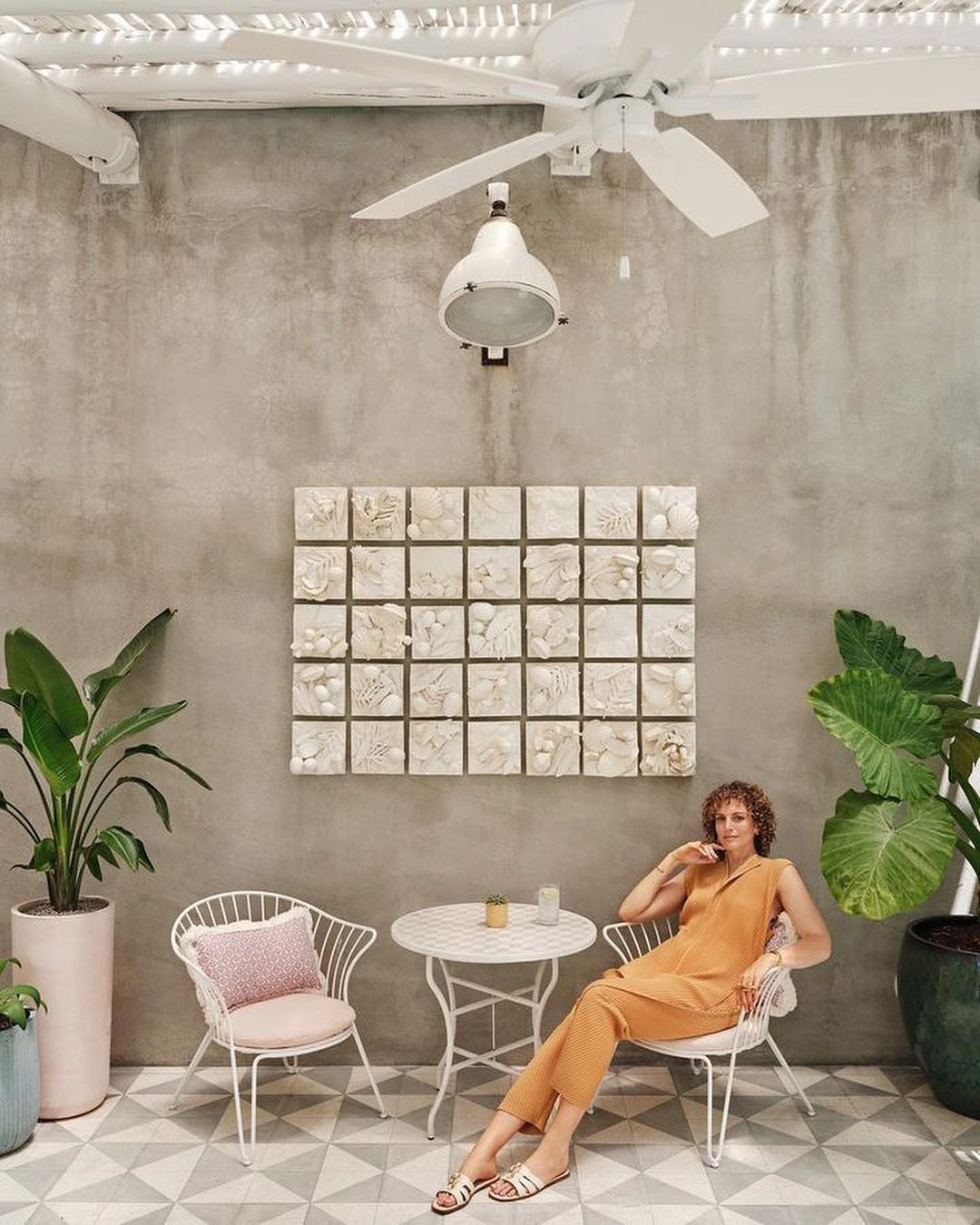 Woman in orange outfit sits at a white metal table on a patio with tile floor, plants, ceiling fan, and wall art.